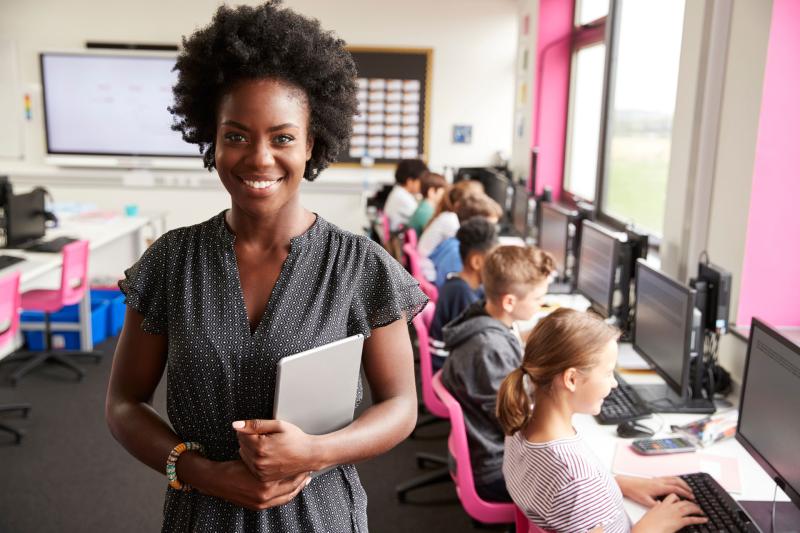 Teacher standing and smiling at the camera while students work on computers in the background.